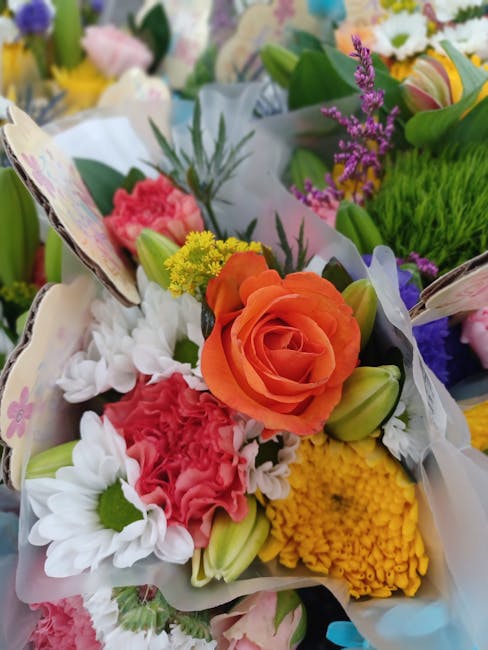 A vibrant bouquet of fresh flowers featuring an orange rose at the center, surrounded by white daisies with green centers, pink carnations, yellow chrysanthemums, and purple veronica flowers. The arra