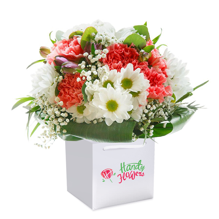 A lush and exuberant floral arrangement stands proudly within a signature white square gift bag, beautifully captured against an immaculate white background. The bouquet shines with soft coral-pink and vibrant red carnations, their velvety, multi-ruffled petals adding texture and a gentle fragrance reminiscent of early summer in Harold Wood's green spaces. Tucked among the carnations are bright white daisies, each encircling a fresh green center that radiates cheerful energy. Sprigs of ethereal baby's breath weave through the composition, lightening the mood with their delicate charms, while a few unopened buds-just kissed with purple-hint at coming blooms and add a whisper of depth. This entire arrangement is ringed by deep, glossy green leaves, forming a living cradle that reflects the beauty of Harold Wood's nature trails. The sturdy white gift bag, topped with elegant ribbon handles, makes for both easy gifting and stylish presentation. Photographed in soft, even lighting, the bouquet's vivid hues and inviting textures are clear and true, marking it as the perfect offering for birthdays, anniversaries, or expressing heartfelt sentiment with local artistry and care.
