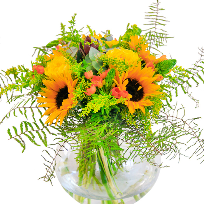 The first glance at this bouquet brings the warmth and radiance of a Harold Wood summer day flooding in. A medley of sunflowers with dark, cocoa-brown centers and beaming yellow-orange petals anchor the arrangement, filling it with lively energy that recalls strolls through local parks or the village green. Close by, yellow garden roses nestle, their petals tightly packed and opening in elegant, buttery spirals, offering softness and depth that complement the bold sunflowers. Scarlet Hypericum berries glint among the green, their shiny skins providing a rich contrast both to the matte finish of the roses and the plush nap of each sunflower. Enveloping the blooms, verdant fern fronds cascade delicately down the sides, mingling with wispy sprigs of light green foliage-possibly goldenrod or solidago, each adorned with tiny yellow blossoms. Bluish, round-leaved eucalyptus peeks out here and there, adding a rustic harmony. All the beauty is captured in a clear, ball-shaped glass vase-like a window into a fresh-cut summer arrangement-where the stems are reflected in cool, shimmering water. The bouquet's scent is light and grassy, subtly floral, conjuring the breeze drifting through Harold Wood's residential lanes. Whether for an anniversary, birthday, or simply to brighten an afternoon, this arrangement is a local classic-crafted with skilled hands and a genuine love of flower artistry.
