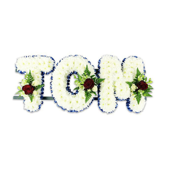A sorrowful and carefully composed floral tribute reads 'TOM' in large, three-dimensional letter shapes, each letter formed by a dense blanket of pristine white chrysanthemum-like blooms whose layered petals create a plush, quilted texture. The entire name rests against a clean white backdrop that amplifies the contrast of colour and detail: a band of royal blue ribbon neatly edges every letter, providing crisp definition and a quietly elegant border that lifts the arrangement's shape. Placed thoughtfully within each letter is a small, symmetrical spray of accent blooms anchored by a single dark red rose-deep, velvety and evocative-surrounded by smaller creamy-white spray roses and tight buds that introduce a softer tonal range. Interplay of textures continues with slender fern fronds and glossy green leaves that fan out to add depth and a natural silhouette, their feathered lines softening the geometric letters. A slender dark-green or black metal framework runs along the base, subtly visible and designed to support the tribute when exhibited at funeral services, memorial events, or in a crematorium chapel near the A12 corridor and local Harold Wood venues. The composition balances formal structure with tender detail: the white chrysanthemums convey quiet consolation while the red roses mark intimate remembrance. As a florist reflecting on the piece, I imagine family members pausing before it-drawn to the tactile softness, the precise ribbon edge, and the single rose that reads like a personal message-an understated but powerful floral homage to a beloved life.