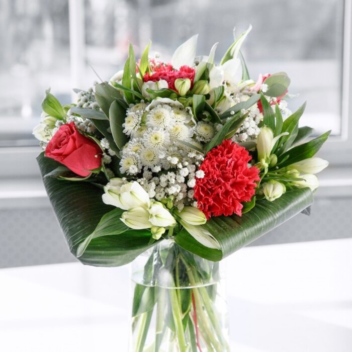 A luxurious red and white floral arrangement stands tall in a clear cylindrical vase, the kind you might find as a centrepiece at a family gathering in Harold Wood, not far from Harold Wood Park. The transparent glass reveals a careful grid of fresh green stems beneath the waterline, each one trimmed cleanly so the bouquet feels structured yet effortlessly natural. Rising from this base, velvety deep red roses create a pulse of colour, their petals gently spiralling open to suggest classic love and devotion. At the front, a large crimson carnation, richly ruffled and full of layered petals, provides a dramatic focal point and a tactile sense of movement. Around these red blooms, soft white flowers introduce balance and calm: elegant white alstroemeria with elongated petals and subtle freckles, rounded chrysanthemum-like florets, and several pristine white lily buds still closed, hinting at future blossoms and a slowly unfolding beauty. Airy clouds of baby's breath weave between the larger heads, their tiny white flowers catching the light and adding a halo of delicacy. The bouquet is framed with lush greenery, including glossy, dark green tropical leaves that curl and wrap around the base like a sophisticated collar, supported by slimmer pointed foliage and silvery eucalyptus-style stems that give depth and gentle variation in tone. The arrangement rests on a smooth white surface with a softly blurred, bright background, as if placed near a sunlit Harold Wood window or patio door. The overall mood is uplifting and inviting, blending modern composure with traditional romance. It would look at home on a dining table off The Drive, celebrating an anniversary, a special birthday, or simply offering an elegant thank you that feels thoughtful and well-crafted.