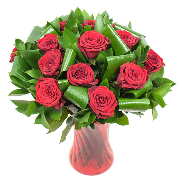 This photograph captures a sumptuous, formal bouquet of deep crimson roses-about a dozen or slightly more-each flower opened to show layered, velvety petals that form tightly spiralled centres, their texture almost tactile beneath smooth, even lighting. The roses are set within a generous matrix of dark green foliage: smaller, glossy leaves provide a dense, shadowed backdrop while larger, bright green leaves are deliberately rolled, curled and folded into elegant loops that cradle the bloom clusters and introduce a sculptural, contemporary silhouette. Those handcrafted leaf forms create visual punctuation around the red heads, balancing the arrangement with modern structural elements while preserving the classic language of romance. The stems sit in a tall, translucent ruby-red glass vase whose subtle ribbing or twisted finish creates vertical highlights that echo the stems' lines and enhance the bouquet's rich colour harmony. With a pure white background, the crimson petals and deep greens stand out crisply, the composition reading as both luxurious and restrained. The image suggests the gentle, rosy scent of fresh-cut roses and the quiet ceremony of giving: ideal for anniversary presents, Valentine tributes or refined centrepieces. Locally, this is the sort of elegant arrangement often chosen by clients in Harold Wood and neighbouring Romford and Gidea Park who want a timeless, expertly crafted statement of love that photographs beautifully and endures in the vase.