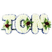 The photograph shows a personalised sympathy arrangement spelling out the name TOM in bold floral lettering, a classic choice for a funeral tribute in Harold Wood. Each of the three letters is constructed in sturdy block form and entirely filled with crisp white chrysanthemums, their countless little petals packed closely together to create a soft, padded appearance that feels gentle to the eye. Framing these white blooms is a royal blue satin ribbon that curves neatly around the edge of each letter, adding a defined outline and a cool, dignified note of colour that hints at formality and respect. At thoughtful positions on the T, O, and M, the white chrysanthemum surface is broken by small, carefully arranged clusters of accent flowers: deep, velvety red roses form the heart of each cluster, framed by smaller white or cream spray roses and slender white flowers similar to freesia. These blooms are nestled into fresh green foliage, including feathery fern and other leaves that provide contrast and a touch of natural movement. The letters sit side by side on a slim, dark support bar, indicating that the tribute is ready to be displayed at a church, crematorium, or graveside in Harold Wood. Set against a clean white background, the textures of petal, ribbon, and leaf are clearly visible, and the arrangement as a whole conveys a blend of purity, love, and remembrance. It feels like a heartfelt way for family and friends to have Tom's name present in flowers as they share memories and say goodbye.