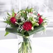 A striking red and white bouquet glows in soft daylight, arranged in a tall, clear cylindrical glass vase set on a crisp white surface, like a dining table in a Harold Wood home. Inside the vase, the freshly cut green stems are neatly aligned and visible through the water, giving a sense of care, craftsmanship, and just-arrived freshness. Rising from this transparent base, velvety deep red roses open in graceful spirals, their petals gently unfurling to suggest timeless romance and heartfelt passion. A large, ruffled crimson carnation sits confidently near the front, its densely layered petals adding rich texture and drama to the arrangement. Around these bold reds, creamy white and pure white blooms provide a gentle contrast: elegant white alstroemeria with slender petals and tiny speckles, plump chrysanthemum and daisy-like centres, and several closed or half-open white lily buds hinting at blossoms yet to come. Wisps of dainty baby's breath are threaded throughout, their tiny white florets like a cloud of lightness, softening the stronger shapes and adding airy volume. The flowers are held and framed by a collar of glossy dark green tropical leaves, some carefully curled around the base, with additional eucalyptus-style sprigs and pointed foliage giving depth and varied shades of green. The background is bright and softly blurred, reminiscent of daylight filtering through a Harold Wood window, making the bouquet feel welcoming and celebratory. The overall mood is sophisticated yet warm, perfect for an anniversary in a home off Gubbins Lane, a birthday surprise delivered to a Harold Wood office, or a thoughtful gesture to brighten someone's day with a classic mix of romance, purity, and joy.