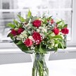 A lush, romantic bouquet stands in a clear glass vase on a bright white tabletop, bathed in soft natural light drifting through a large window, like one overlooking the quiet streets of Harold Wood on a calm morning. Deep crimson red roses, some fully open with velvety petals spiralling outward and others still held in tight, elegant buds, form the passionate heart of the arrangement. Around them, rich red carnations with densely ruffled petals add fullness and texture, echoing the classic charm of traditional English floral design. Nestled between the bold reds are crisp white alstroemeria, or Peruvian lilies, many still in bud with green-tipped petals, while a few partially open blooms reveal delicate, trumpet-shaped flowers with subtle markings. Tiny white gypsophila, or baby's breath, is woven throughout like a soft, cloudlike mist, gently lightening the deep reds and giving the bouquet an airy, ethereal quality. A generous collar of glossy, broad green leaves cradles the base of the flowers, while finer, lighter green foliage threads through the design, adding movement and layered texture. The stems, neatly trimmed, are clearly visible through the transparent glass, immersed in clean water that catches a faint reflection from the pale surface below. The background scene beyond the window is softly blurred and nearly monochrome, suggesting an indistinct garden or street near Harold Wood station, which makes the vivid reds, whites, and greens appear even more intense and alive. The overall mood is one of heartfelt celebration and refined romance, ideal for local birthdays, anniversaries, or a same-day delivery gesture to brighten a home along Gubbins Lane or near Harold Wood Park.