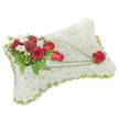 A quiet, reverent sympathy pillow formed from a dense carpet of small, snow-white chrysanthemums fills the image, their tight, velvety heads creating a plush, cloud-like surface that suggests rest and peace. Around the slightly raised edges a thin band of pale green ribbon or foliage peeks through, framing the rectangular cushion with a subtle freshness that feels like a gentle breath of spring. On the upper-left corner an artful spray of deep red roses and rich burgundy carnations sits with poised dignity; their velvety petals and layered ruffles bring warmth and a tender intensity against the pure white base. Sprigs of tiny white accent blooms, likely freesias or small spray chrysanthemums, lift lightness and fragrance into the composition while feathery fern fronds and glossy leaves form a calming green backdrop. From that main cluster, slender arcs of bright green bear-grass or similar foliage sweep diagonally across the chrysanthemum field toward the lower right, creating a graceful line of movement that carries the eye and the sentiment across the piece. The trail culminates in a single, perfectly formed red rose resting on the bottom right of the pillow, a clear symbol of enduring love and remembrance. Soft, neutral studio light highlights the textures and the gentle contrast between the white cushion and the saturated reds, evoking the quiet calm of a chapel or a bedside in Harold Wood on a still morning. This classic memorial arrangement, both simple and carefully detailed, reads as an expression of comfort and respect, suitable for funeral services, wakes, or a heartfelt tribute left near the family at a local church or Harold Wood Station memorial.