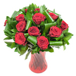 A lush, rounded bouquet of roughly a dozen deep crimson roses sits at the heart of this image, each bloom opened to reveal velvety spirals of tightly layered petals that catch soft, even light and give the flowers a satin, wine-dark sheen. The roses are nestled in a dense collar of glossy, dark green foliage - smaller ruscus- or salal-like leaves forming a verdant backdrop while larger, bright green aspidistra-style leaves are expertly curled and looped into sculptural spirals that frame and cradle each head. Those folded leaves add a modern, architectural rhythm against the gentle curves of the roses, creating depth and movement across the composition. The stems are held in a tall, translucent ruby-red glass vase with a subtle ribbed or twisted surface; vertical lines in the glass pick up highlights and echo the vertical flow of the arrangement, harmonising with the passionate red of the petals. Set against a clean white background, the deep reds and rich greens pop with dramatic clarity, and the image feels both intimate and luxurious. There is an implied scent of classic rose perfume-warm, slightly honeyed-suggesting this bouquet would be a considered romantic gift for an anniversary or Valentine's Day, or a sophisticated centrepiece for a candlelit dinner. As a local floral note, this is the kind of arrangement our customers in Harold Wood and nearby Romford request when they want timeless elegance with a contemporary twist, presented with careful craftsmanship and fresh, long-lasting stems.
