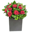 A modern, sculptural bouquet of deep crimson roses sits arranged like a small chorus in a low matte black cube - each bloom velvety and full, petals unfurling in soft, generous spirals that catch the light with a subtle, satin sheen. Around the roses, broad glossy green leaves have been carefully curled, folded and layered to create rhythmic waves of texture and movement; the foliage feels almost architectural, its polished surfaces reflecting highlights and adding depth behind and between each crimson head. The square box vase gives the design a grounded, contemporary silhouette, the matte finish quiet against the vivid richness above so the roses become the unmistakable focal point. The lighting is clear and modern, a steady brightness that picks out the velvet-red tones and the gleam on every leaf so the whole composition reads as both intimate and confidently styled. If you bring the bouquet close you can almost imagine the scent: the classic, heady sweetness of rose softened by refreshingly green notes from the leaves - a clean, uplifting perfume that suits a modern home or office. As a local florist I see this as a deliberate statement piece, made to mark anniversaries, romantic milestones, or to elevate a simple table with considered drama. It carries the quiet confidence of careful craftsmanship - an urban arrangement with the warmth of thoughtful intent, fitting for a living room in Harold Wood or a bedside gift picked up after a walk from the station.